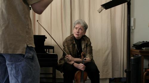 莎莉·托马斯手里拿着小提琴和弓坐着, 抬头看着一个站在画面外的学生. 托马斯的面部表情是一种关注，也许是询问或讨论. 这个房间的装饰简单实用, 有一个普通的幕布背景和一个音乐架, 这说明他很注重音乐练习.